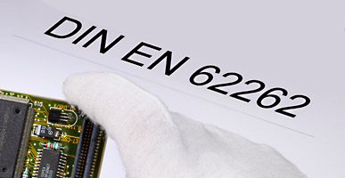 Auf einem Dokument steht DIN EN 62262 geschrieben, darüber ist der Ausschnitt einer Elektronikplatine zu sehen.