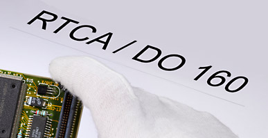 Auf einem Dokument steht RTCA/DO 160 geschrieben, darüber ist der Ausschnitt einer Elektronikplatine zu sehen.