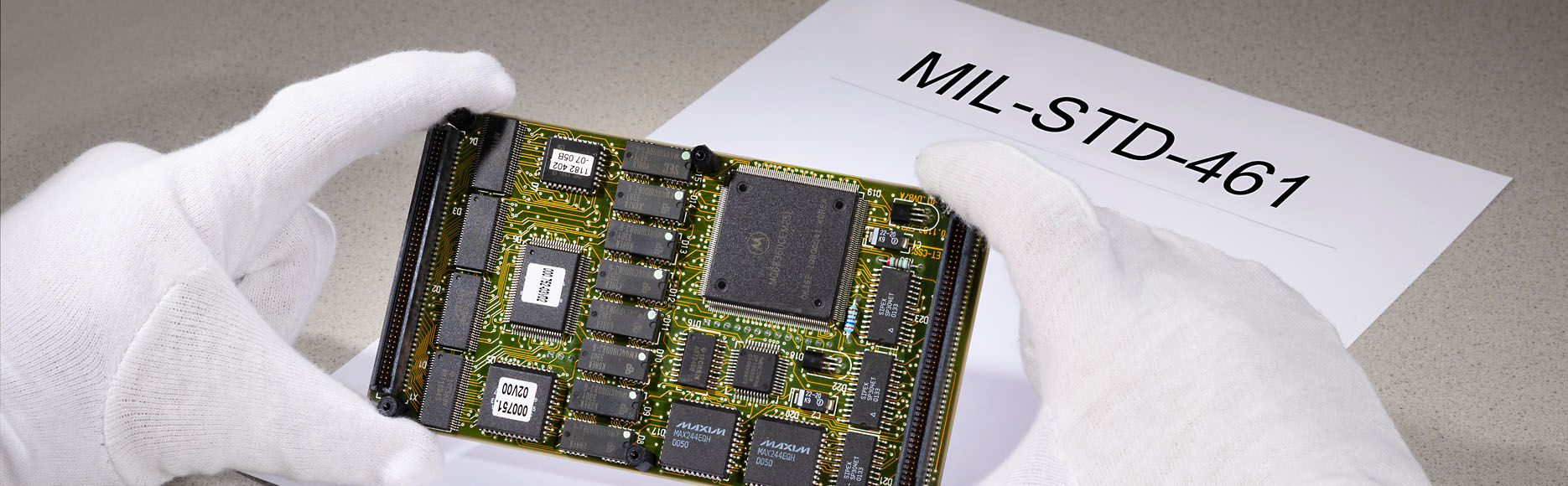 Person wearing gloves holding circuit board in hand. “MIL-STD-461” can be read printed on a document