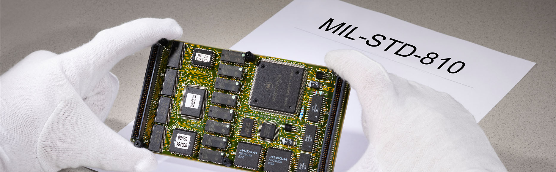 Person wearing gloves holding circuit board in hand. “MIL-STD-810” can be read printed on a document
